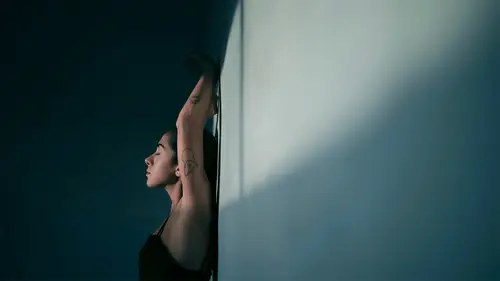 Frau mit Tattoos streckt sich an einer Wand, die Augen geschlossen, im sanften Licht.