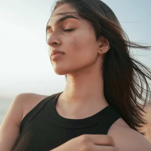 Eine Frau mit langen Haaren steht im Freien, die Augen geschlossen, in einem schwarzen Oberteil. Der Wind weht ihr Haar vor einem klaren Himmel.